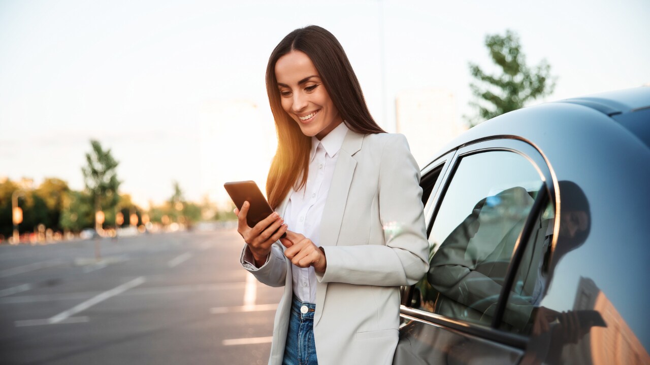 Frau lächelt und blickt auf ihr Handy, während sie vor einem Auto steht.