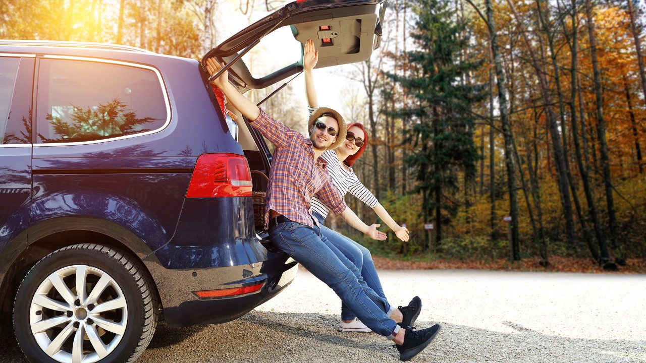 2 Reisende sitzen auf der Rückseite eines Minivans mit weit ausgebreiteten Armen und lächelnd.